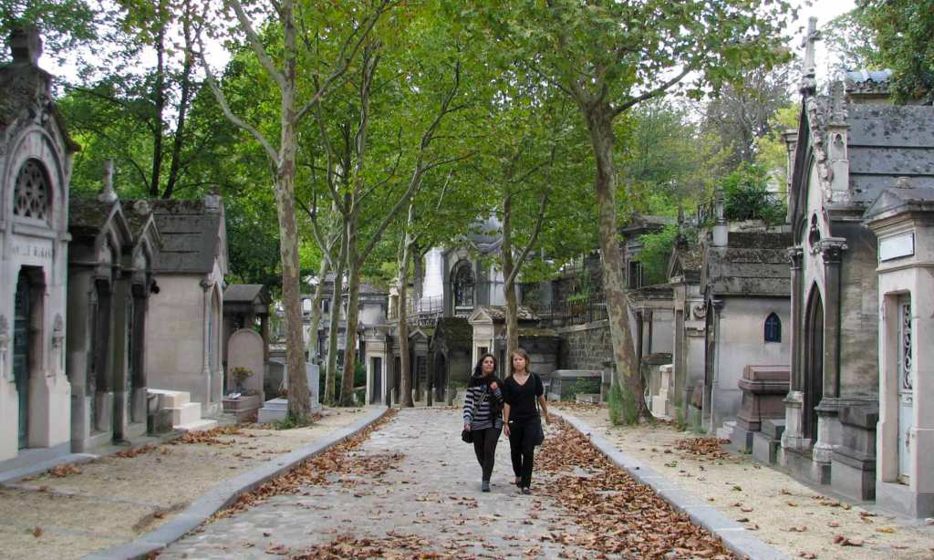 Two women stroll along a lane in Paris' Père Lachaise Cemetery. On either side of the lane there are grey mauseoleums. The street is partially covered with brown autumn leaves.