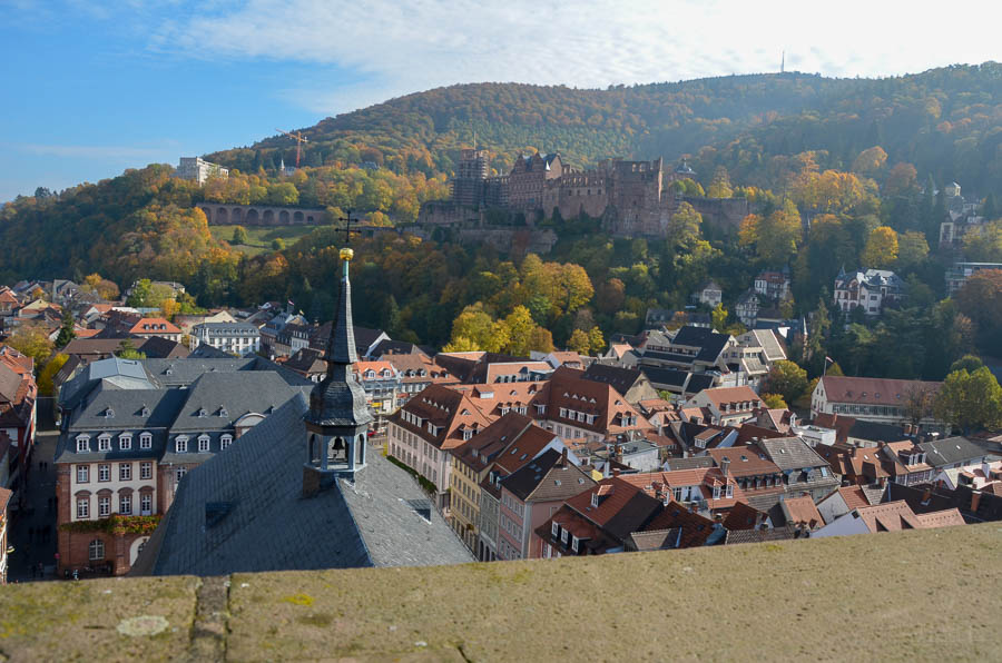 The Heidelberg Castle sits on a hillside filled with trees wearing autumn leaves. The terracotta rooftops of Heidelberg's Old Town buildings are clustered below.