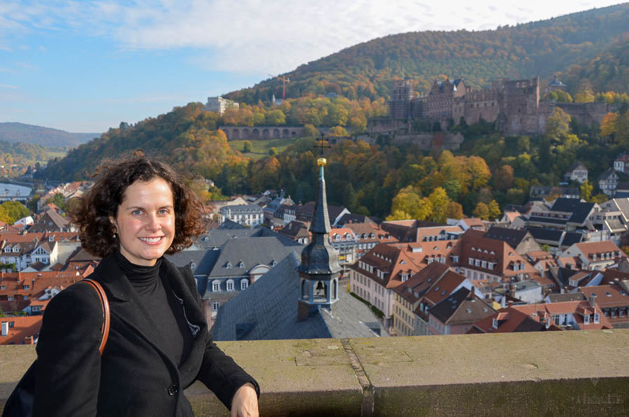 A woman stands on the viewing platform of Heidelberg's Heiliggeistkirche on an autumn day with the Heidelberg Castle visible on the hill in the background, along with autumn leaves. 