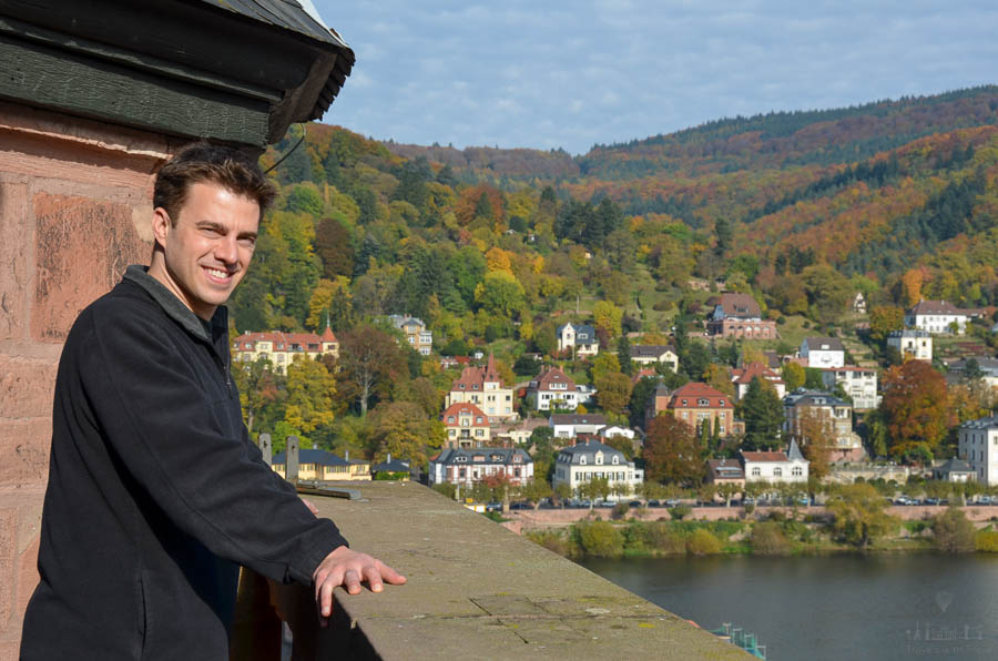 A man stands on the viewing platform of Heidelberg's Heiliggeistkirche on an autumn day. Buildings are visible in the background along with the Neckar River and autumn leaves.