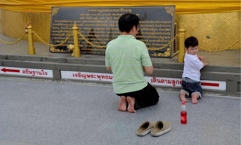 As the adult next to him prays at Bangkok's Golden Mount, a young boy is distracted by a bottle of soda.