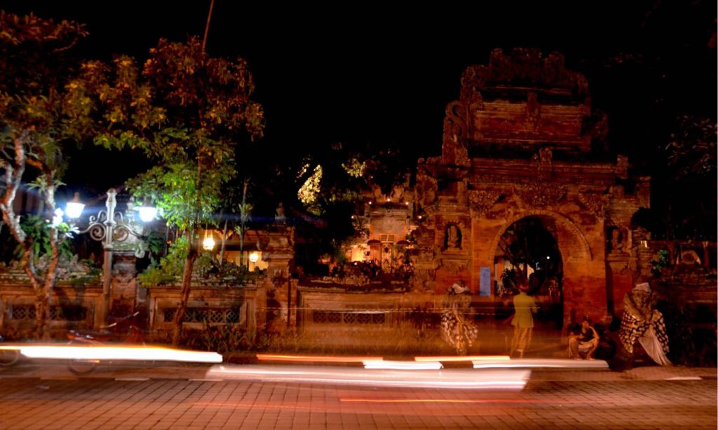 Motorcycles and cars drive past Udud's Royal Palace at night.