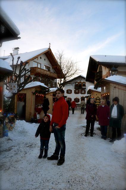 People stand outside a Christmas market in Oberammergau, Germany. There is snow on the ground.