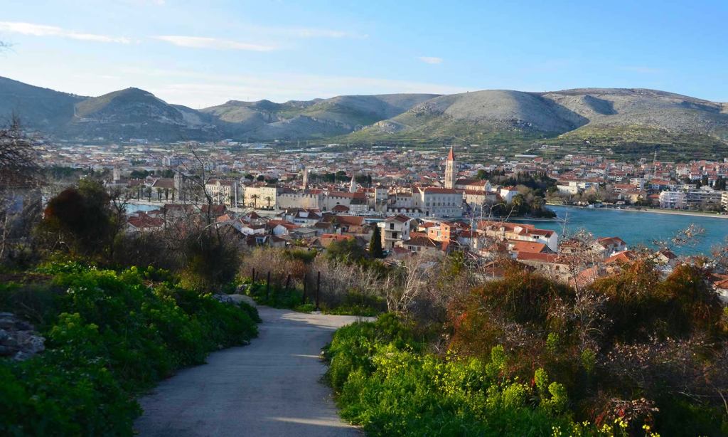 The city of Trogir, Croatia is seen from afar, via a walking path that is surrounded by greenery. You can see mountains in the distance, as well as the Adriatic Sea.
