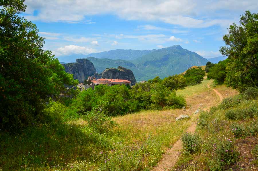 A dirt path winds through the grassy field with the terracotta roof of a Meteora monastery and mountains off in the distance.