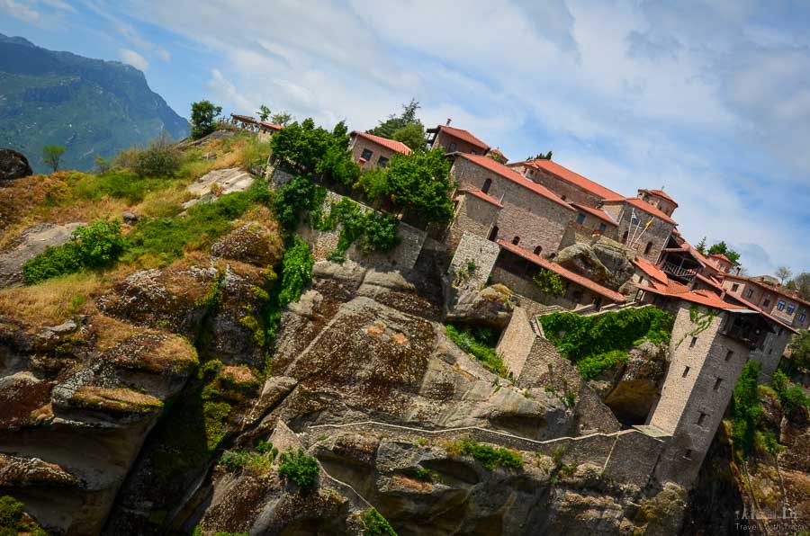 The Great Meteoron Monastery sits on top of a steep mountain, partially dotted with vegetation, in Meteora, Greece.