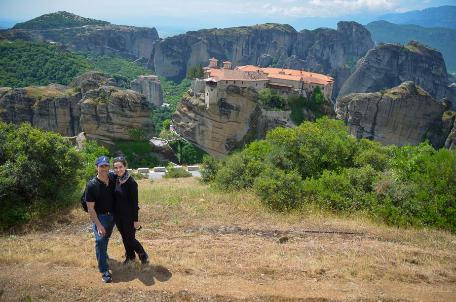 A man and woman stand on a hillside with a monastery in Meteora, Greece and rock formations behind them during a hiking tour. 