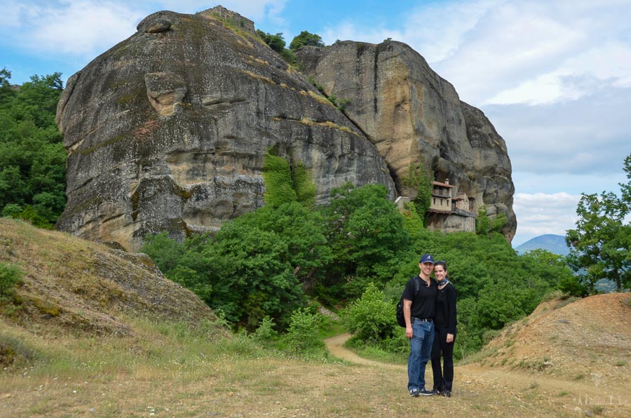 A man and woman on a hiking tour in Meteora, Greece pause on a path, with the rock formations and the Ypapanti Monastery behind them.