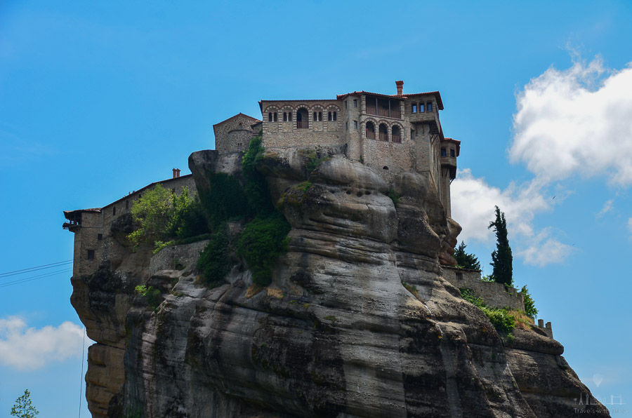 The Holy Monastery of Varlaam sits on a dramatic rock formation on a blue-sky day in Meteora, Greece.