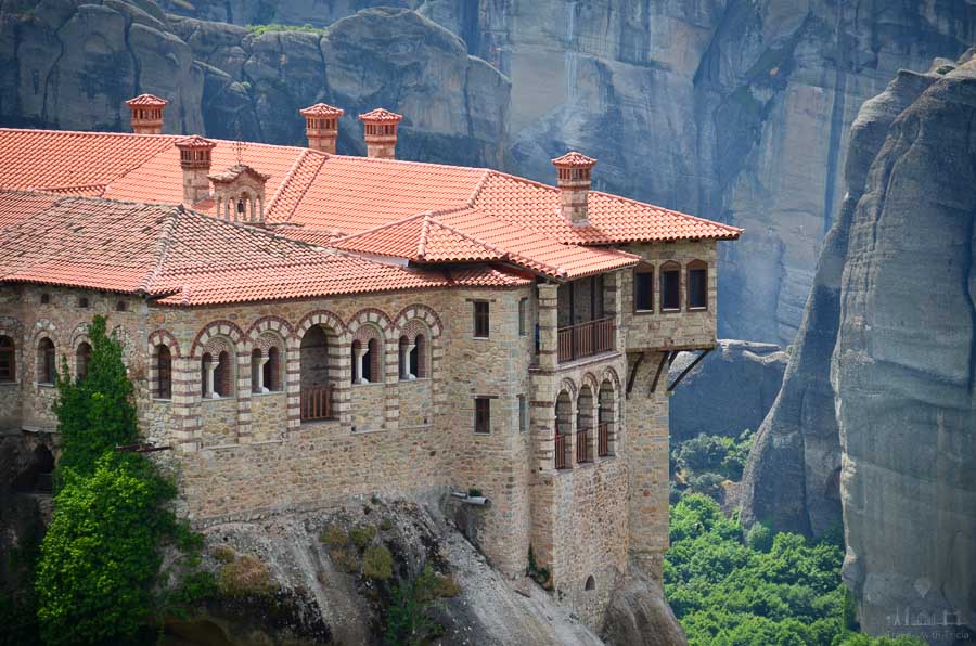 The Varlaam Monastery sits on a steep rock with steep rock formations in the background and green foliage in the valley floor below.