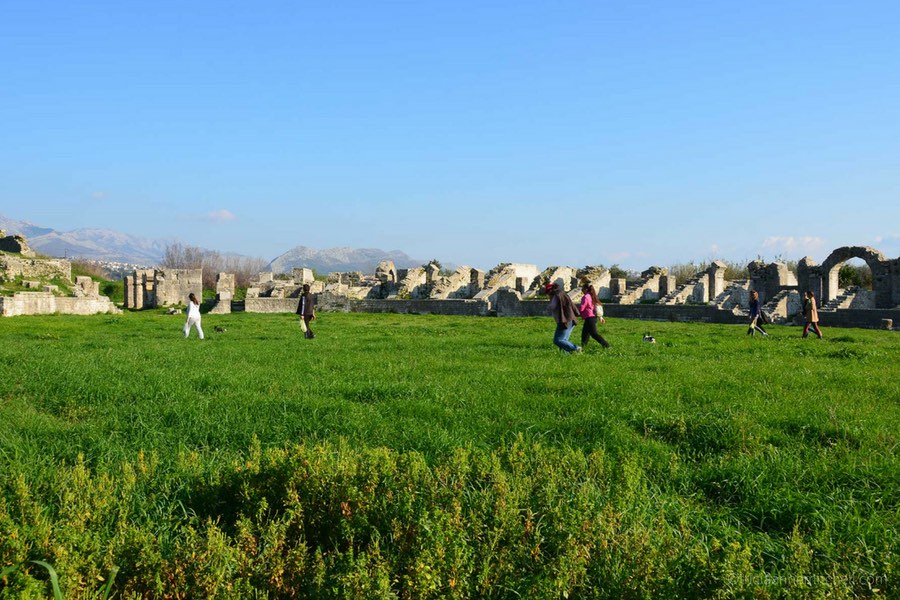 Several people walk through a grassy field, which is encircled by the ruins of Salona's ancient Roman amphitheater.