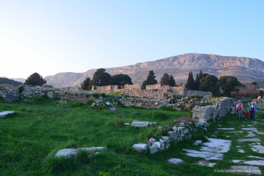 A family walks on one of Salona's weathered stone roads just before sunset. Rugged mountains overlook the scene.