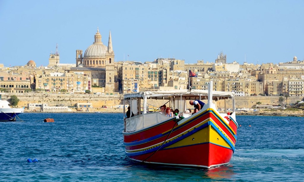 A multi-colored wooden fishing boats sits in the water, with the skyline of Valletta, Malta in the background.