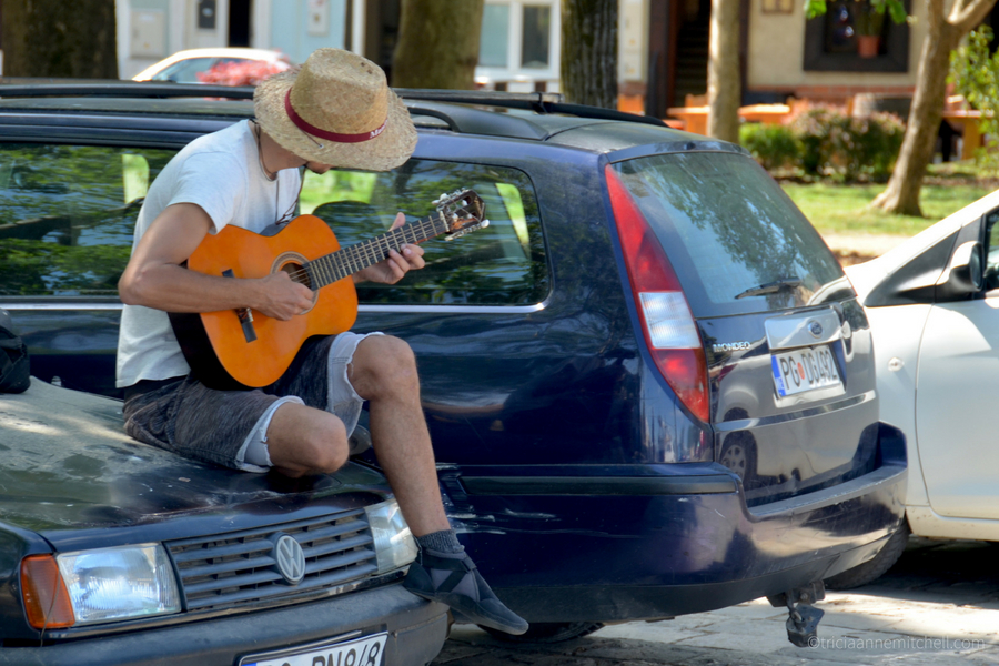 A man sitting on the front of a car plays guitar in a Virpazar parking lot, near Skadar Lake, Montenegro.