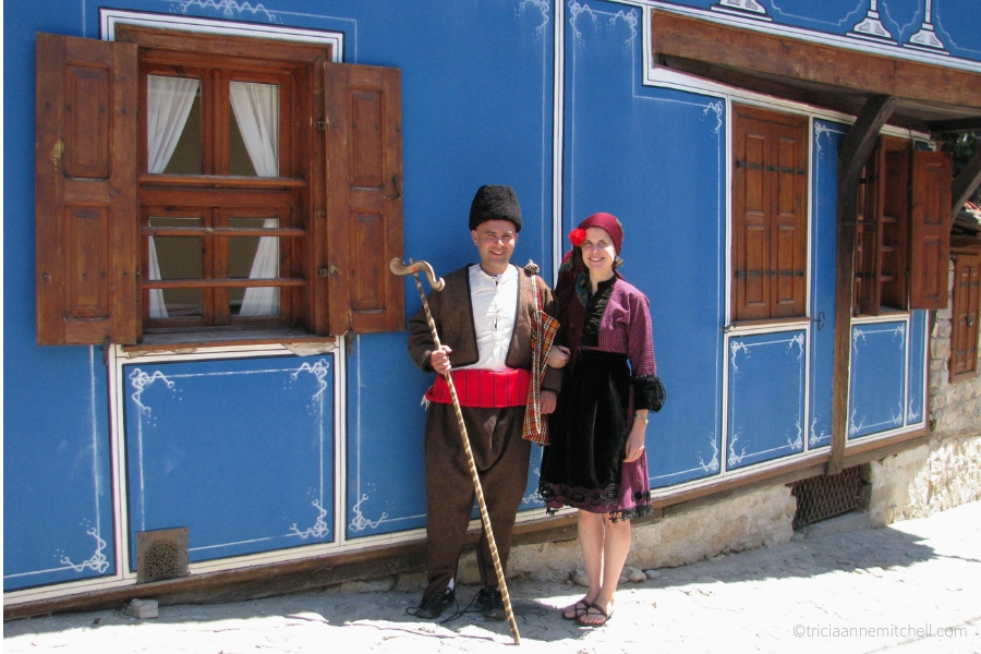 A man and woman, dressed in traditional Bulgarian clothing, stand next to a historic blue building in Koprivshtitsa, Bulgaria.