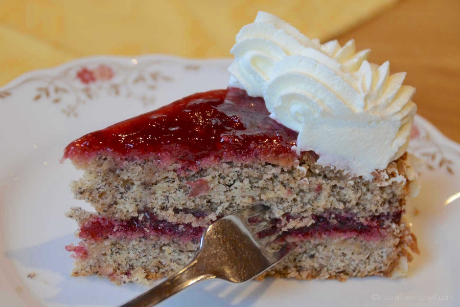 A fork cuts through a slice of layered cake. The flour is golden brown, the jelly-like frosting is red, and there is a dollop of piped-on whipped cream.