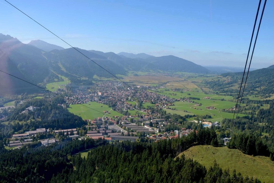 An aerial view of Oberammergau, Germany. The rooftops of buildings are visible, as are evergreen trees and mountain slopes. The sky is blue, and two cable car wires are visible, as the photo was taken from inside the gondola.