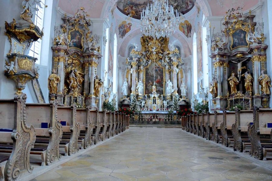 The ornate interior of Oberammergau's church is pictured: You can see old wooden pews, which are heavily carved. There is also a crystal chandelier, ceilings painted in a pale pink, and saints' statues, and many gold accents.