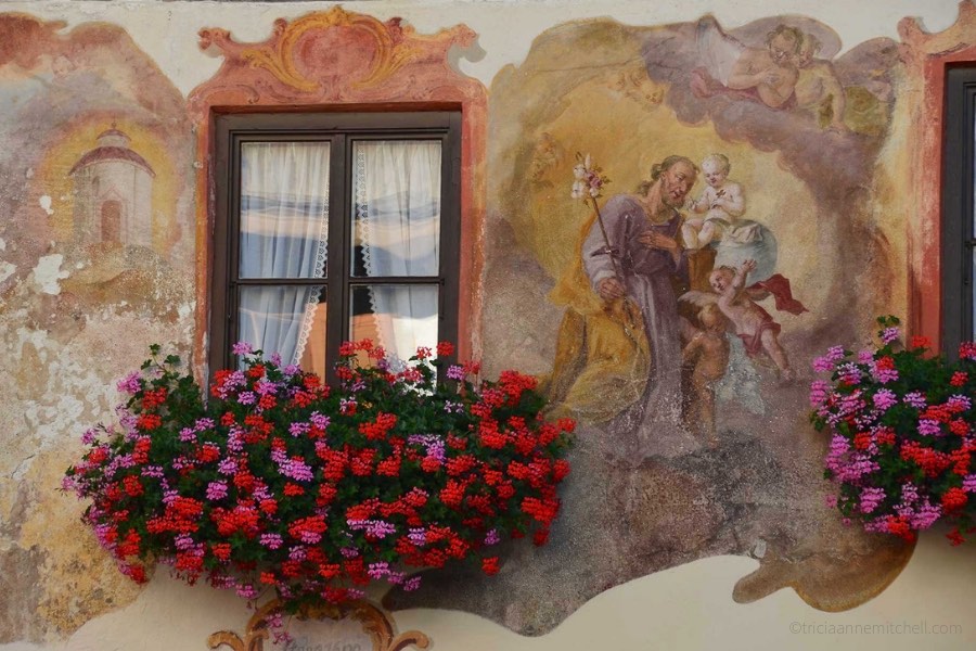 Detail of a home in Oberammergau, Germany: Red and pink flowers fill a flowerbox, alongside a religious mural.
