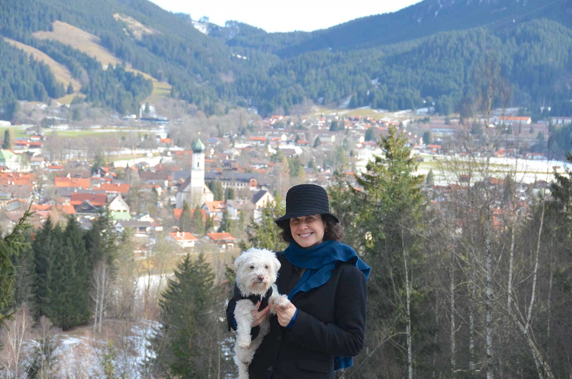 A woman holding a white dog stands outside in nature. The village of Oberammergau is visible behind her, as are forested mountain slopes.