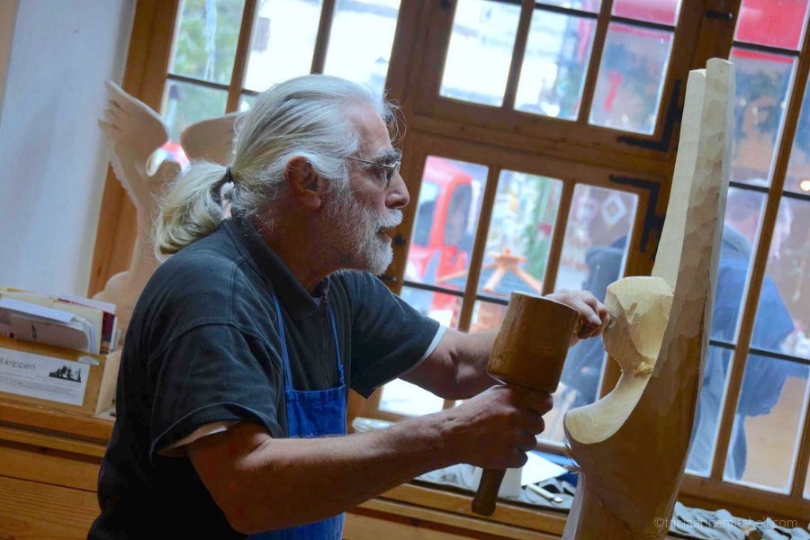 A man with a bear carves an angel out of a block of wood. He is working in front of a window, inside a workshop in Oberammergau, Germany.