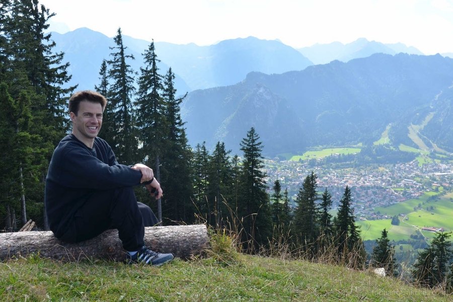 A man sits on top of a mountain slope, overlooking the village of Oberammergau, Germany below.