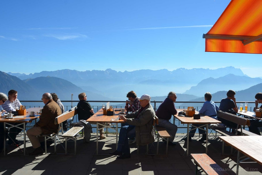Sitting at tables on an outside terrace of the Laber Mountain's restaurant, people look out at the panoramic views. The sky is blue, and the corner of an orange umbrella is visible.
