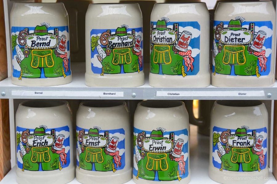 Eight stoneware beer mugs sit on shelves, for sale in an Oberammergau, Germany souvenir shop. Green leather shorts, pretzels, and a Bavarian hat are