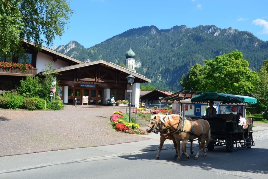 A carriage, pulled by two light-brown horses, drives in front of Oberammergau's Tourist Office and the Ammergauer Haus.