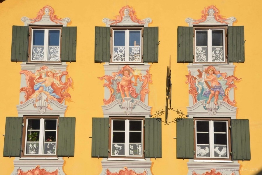 A close-up of a traditional Bavarian home: The wall is painted yellow, there are 6 green shutters, and murals of angels.