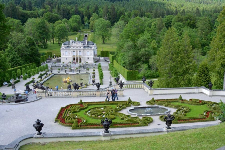 An overhead view of Linderhof Palace's terraced gardens and fountains. The hills surrounding the castle are covered in green trees.