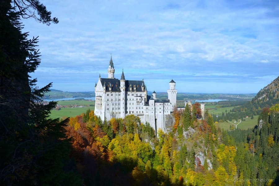 Germany's Neuschwanstein Castle, surrounded by red, green, and yellow foliage, on a sunny day.