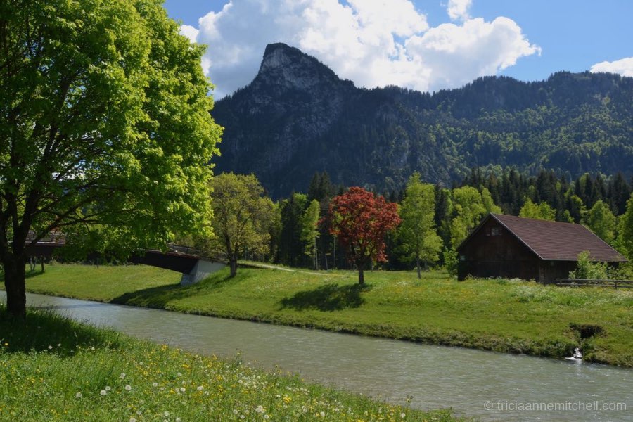 The Kofel, a cone-shaped mountain towers over a river and wooden barn. Green trees are visible.