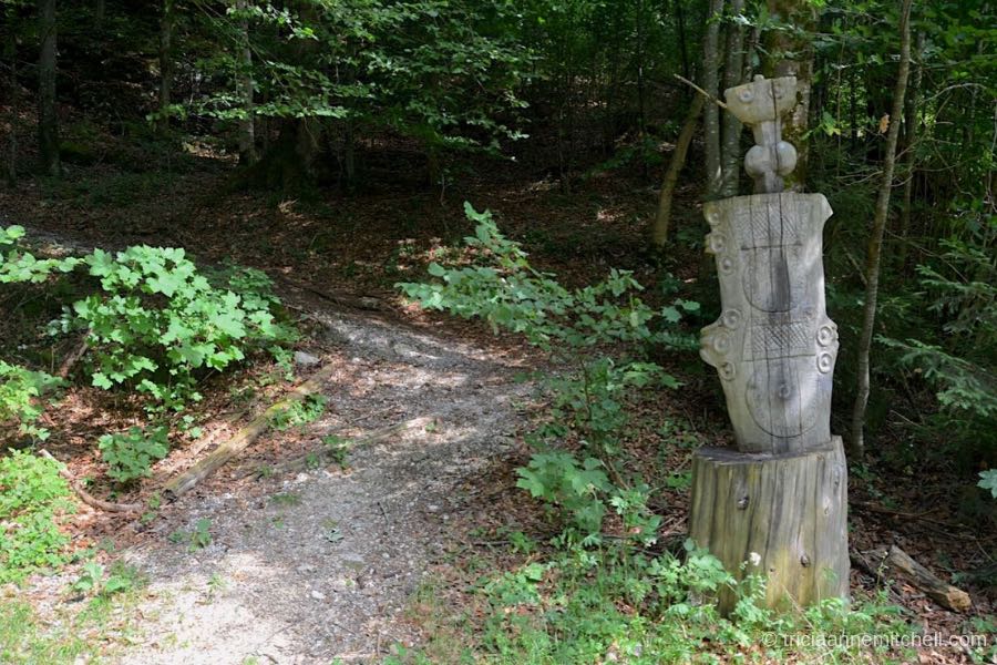 A crushed gravel path leads up a forested hill, past a tree stump that has been carved to look like an ancient Roman dagger.