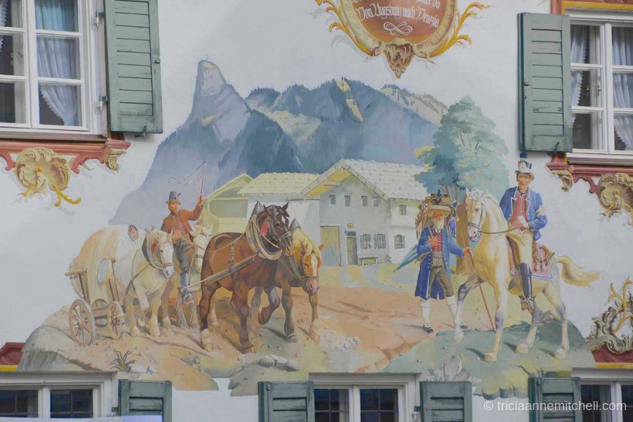Detail of a mural in Oberammergau, Germany: Horses pull a cart, alongside a merchant carrying wooden objects on his back for sale. A mountain and Bavarian-style home are visible in the background.