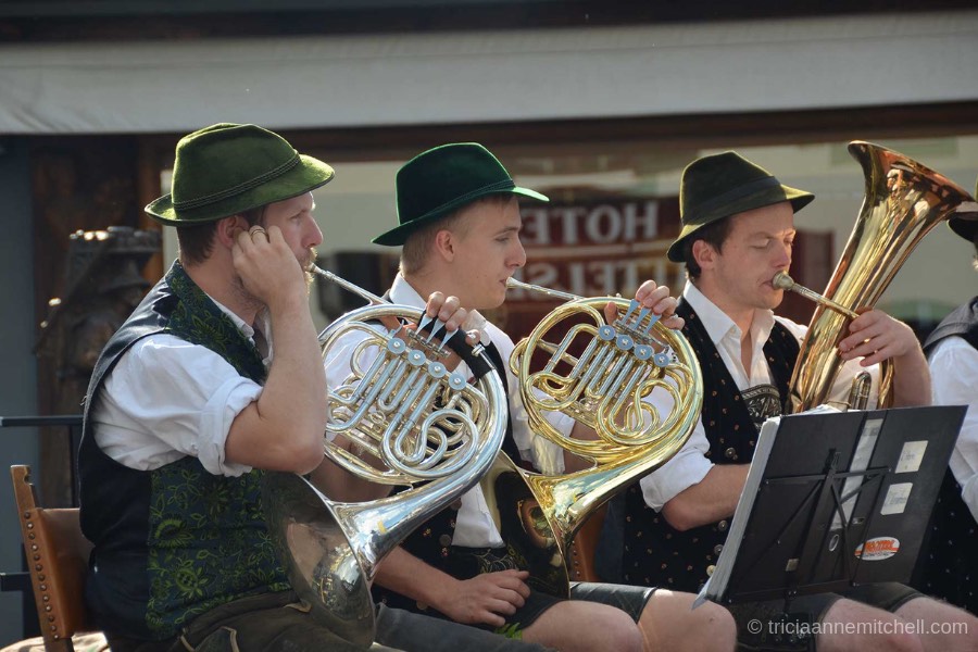 Three men play brass instruments, including French horns, at a festival in Oberammergau, Germany. The men are wearing green Bavarian hats, and traditional vests.