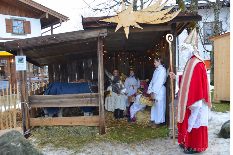 A donkey, girls dressed as angels, and an adult dressed in a red robe of Saint Nicholas stand inside a wooden shed in Oberammergau, Germany. Straw and snow are visible on the ground.