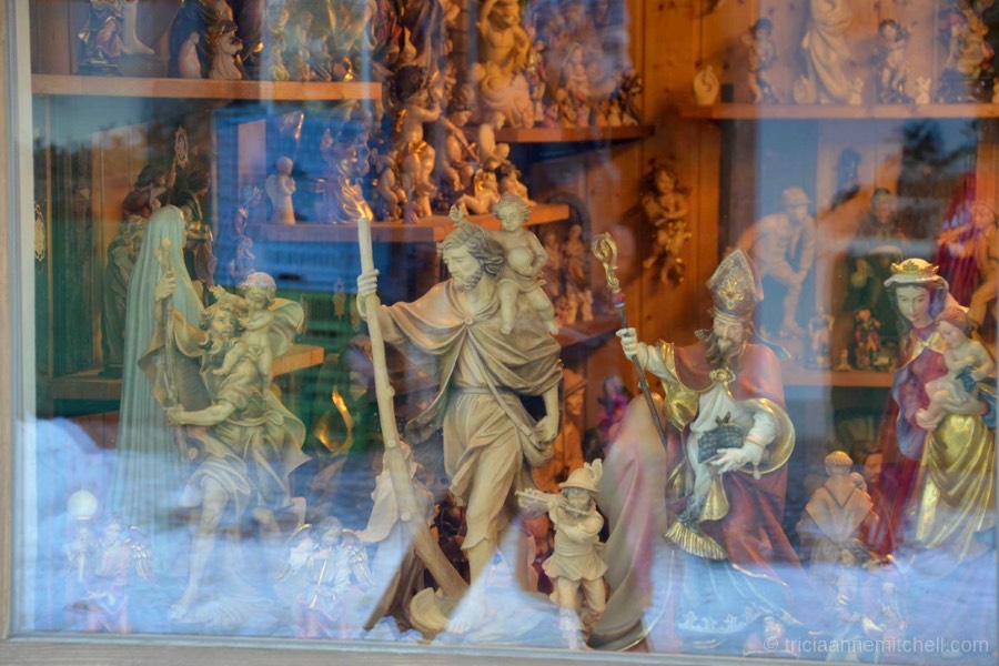 A variety of woodcarvings depicting people and religious saints are visible, viewed through a shopkeeper's window in Oberammergau, Germany.
