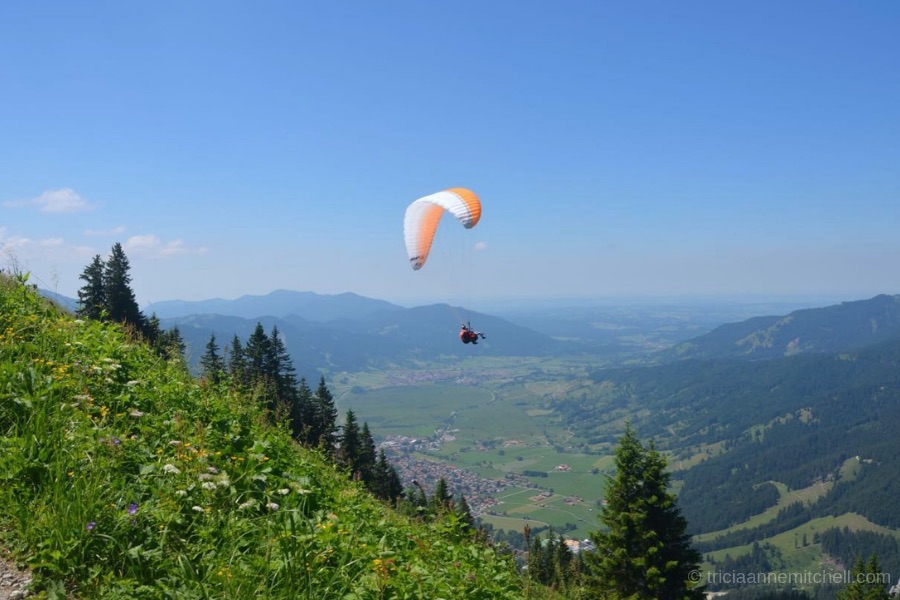 A paraglider with an orange and white parachute flies over the town of Oberammergau, Germany. A steep green hillside carpeted in grass and flowers frames one side. On the other side, you can see the Aufacker Mountain.