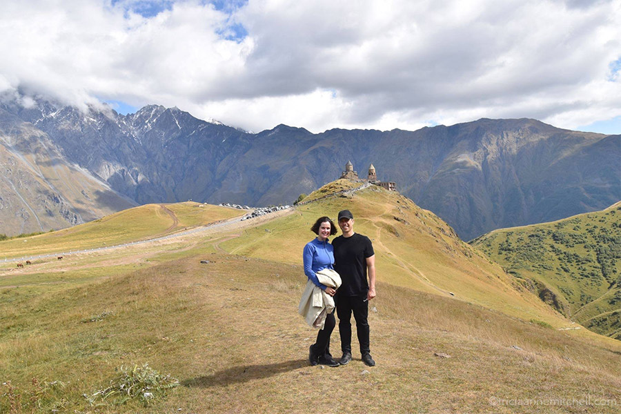 A man and a woman dressed in hiking attire stand in front of the Gergeti Trinity Church in Kazbegi / Stepantsminda, Georgia.