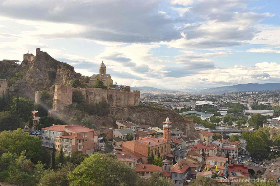 The Narikala Fortress sits on a rocky ridge, towering over red-roofed buildings in the city of Tbilisi, Georgia. The futuruistic Peace Bridge is off in the distance.