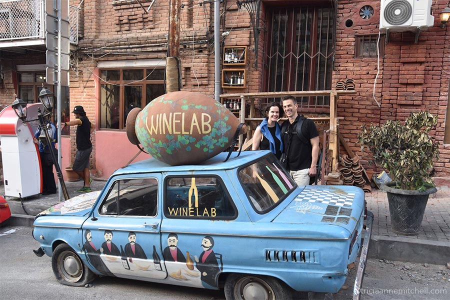 A man and a woman stand next to a blue vintage car parked on a street in Tbilisi, Georgia. On top of the car's roof is a terracotta qvevri, with the name "WINELAB" written on it.