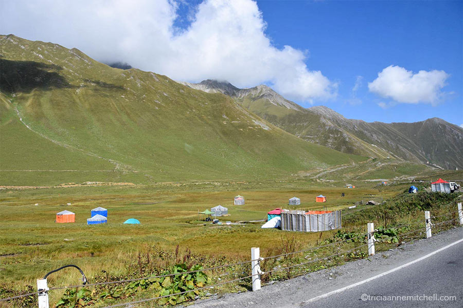About 7 yurts (ranging in color from blue to orange) dot green mountainous slopes alongside the Georgian Military Highway.