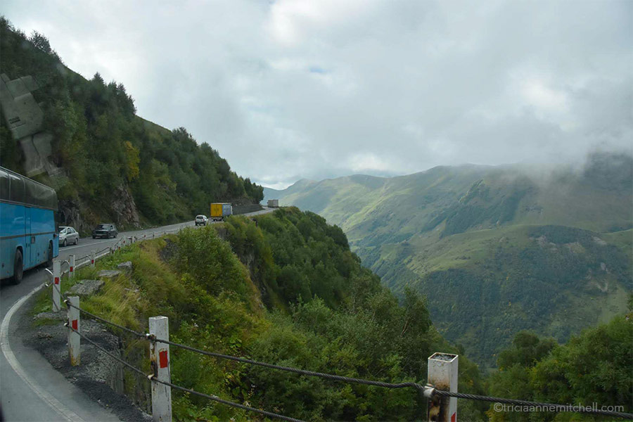 Cars drive on a curvy mountainous highway in Georgia. There is a wire cable guardrail, and steep, green-covered cliffs on the right.