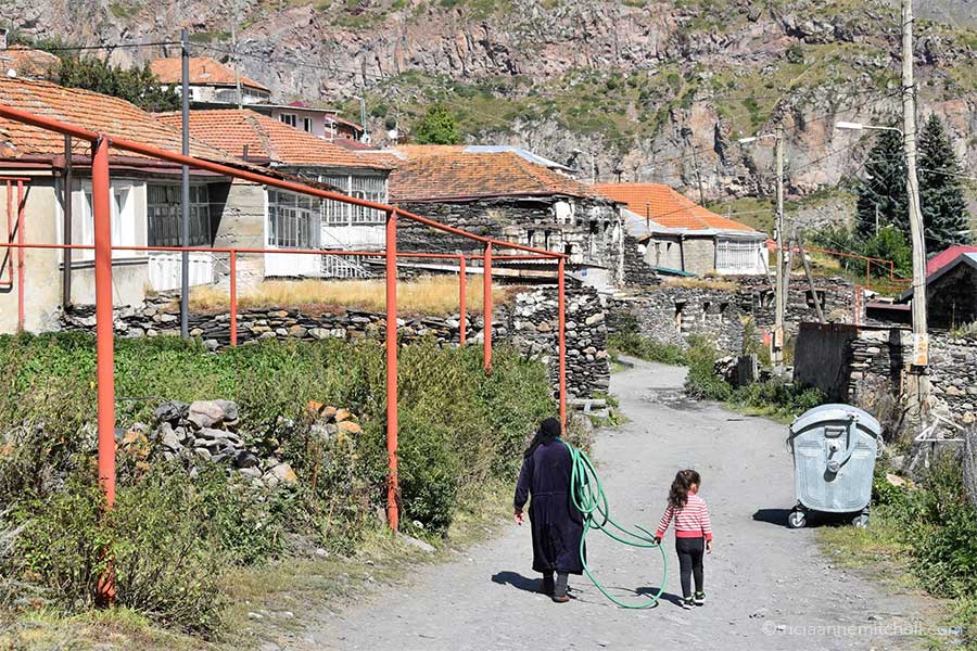 An older woman dressed in black carries a green hose with the help of a young girl. They are walking in a Georgian village with stone houses.
