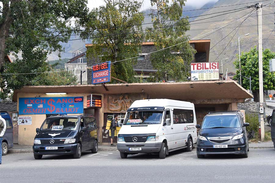 Three minivans are parked by Kazbegi's taxi stand / marshrutka meeting point. The sign on the left indicates money exchange and says "RUB GEL Exchange." Another sign on the right shows the Tbilisi marshrutka schedule. It reads: "Tbilisi Marshrutka, 10 Gel. 7:00, 8:00, 9:00, 10:00, 11:00. 12:00, 13:00, 14:00, 15:00, 17:00, 18:00."