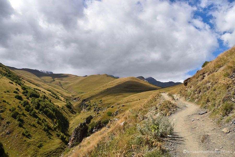The dirt trail leading to the Gergeti Trinity Church in Kazbegi Georgia. The surrounding hillsides are covered with a yellowish-green grass, and the sky is mostly covered in clouds.