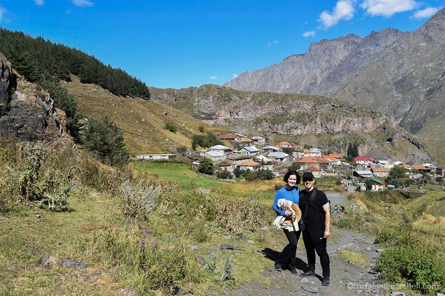 A woman and a man stand among mountain scenery in the Republic of Georgia. A cluster of houses are visible in the background, as are rugged mountain slopes.