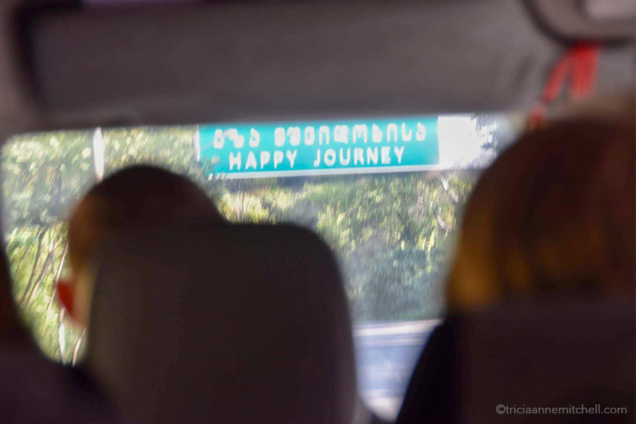 A street sign reading "Happy Journey" in English and in Georgian, is visible through the windshield of a minivan.