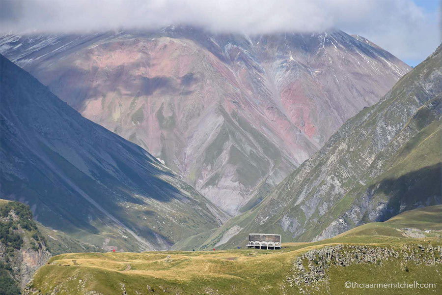 The circular Russia–Georgia Friendship Monument rises from a green plateau. It is surrounded by steep greenish-reddish slopes and is located on the Georgian Military Highway.